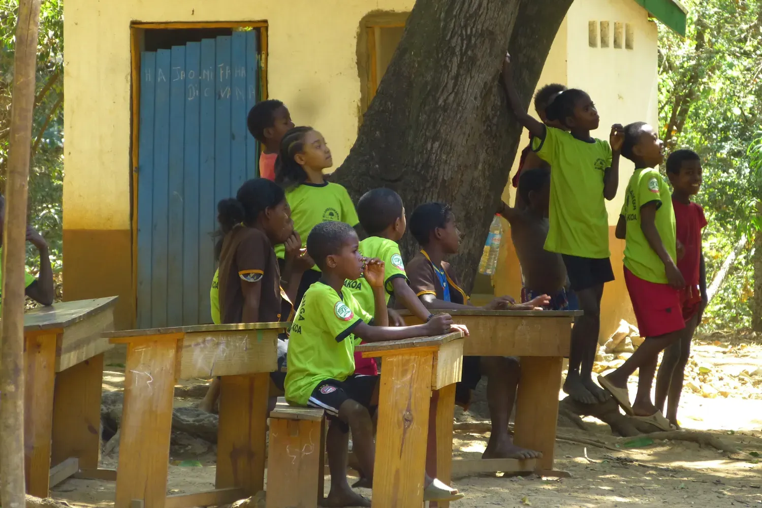 Environmental education in action at the “Green Saturday School” Children in green and brown school uniforms gather around desks and trees in an outdoor classroom setting.