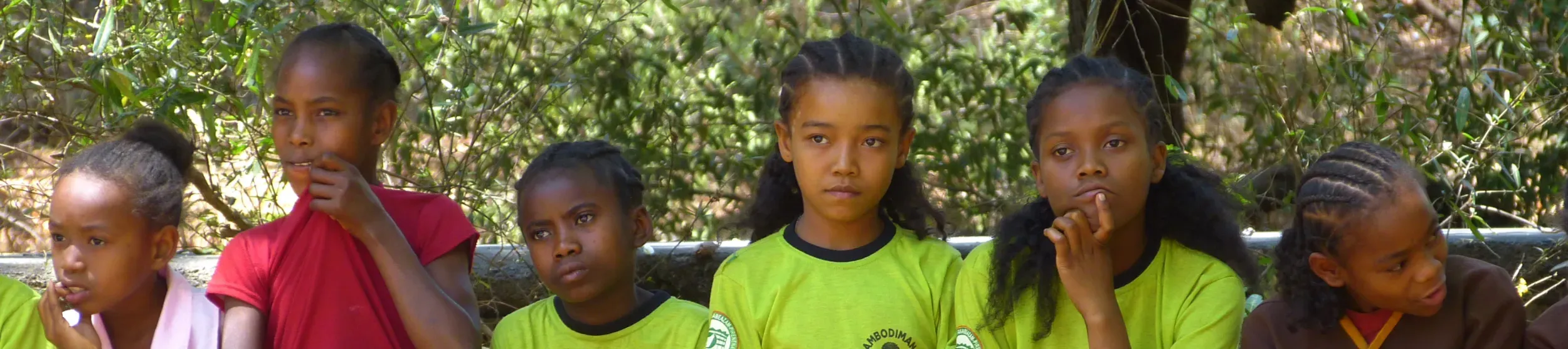 Students from Ambodimanga, a village near Ankarafantsika National Park A group of schoolchildren sit on a wooden bench outdoors, some wearing green uniforms with the school name and logo.